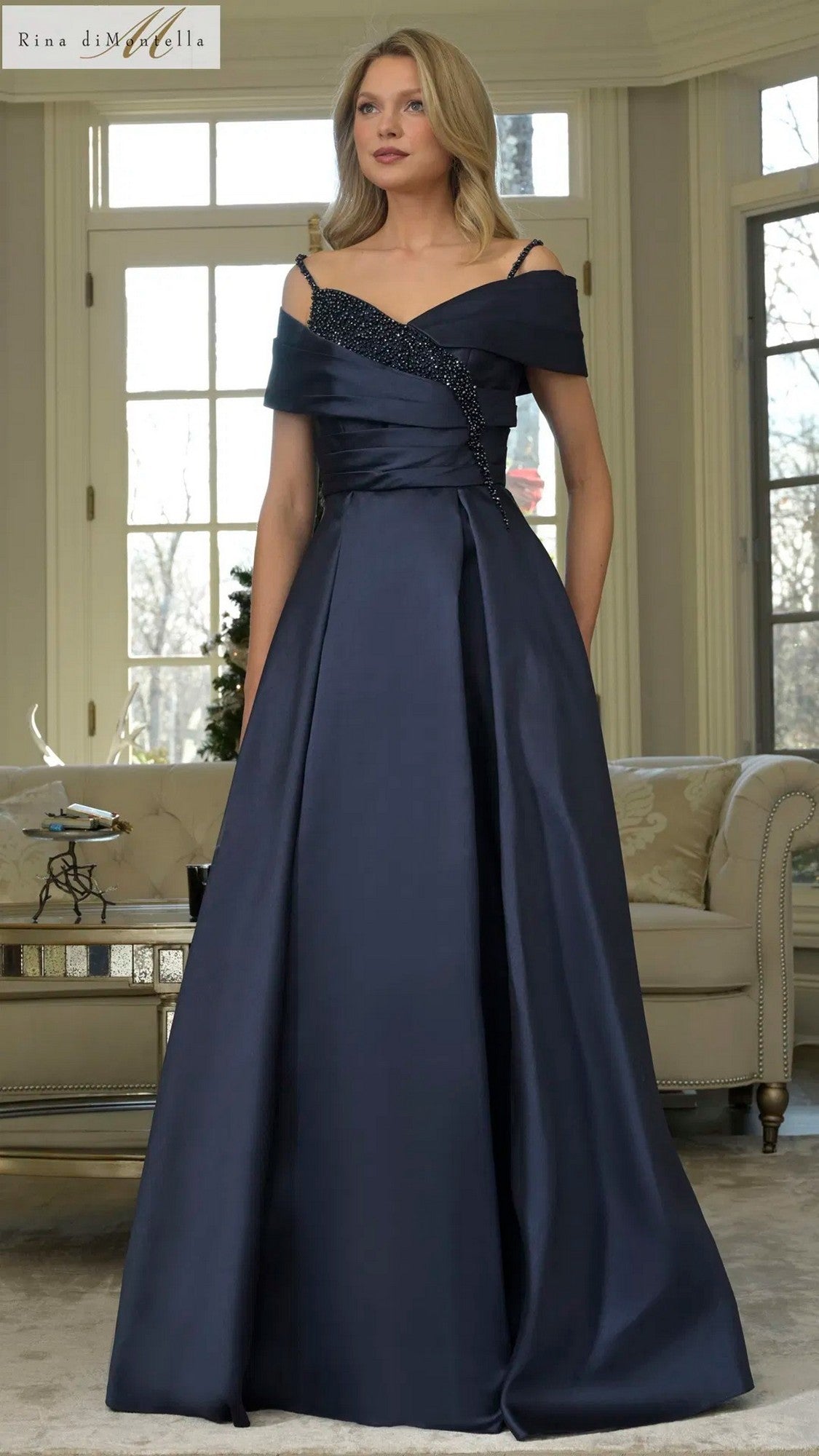 A woman stands indoors wearing the Rina Di Montella RD3221, a navy long formal dress with an off-shoulder beaded bodice and a full flowing skirt.